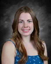 A young woman with long brown hair smiles in a blue dress with white polka dots against a dark background.