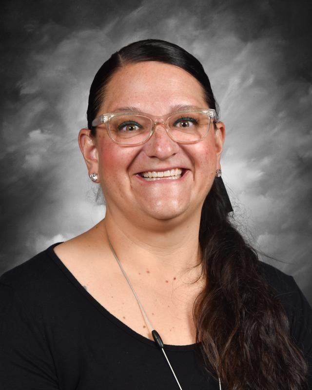 Smiling person with glasses and long dark hair in a ponytail, wearing a black top, against a gray background.