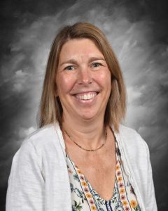 Smiling woman with shoulder-length hair, wearing a floral top and white cardigan, against a gray background.
