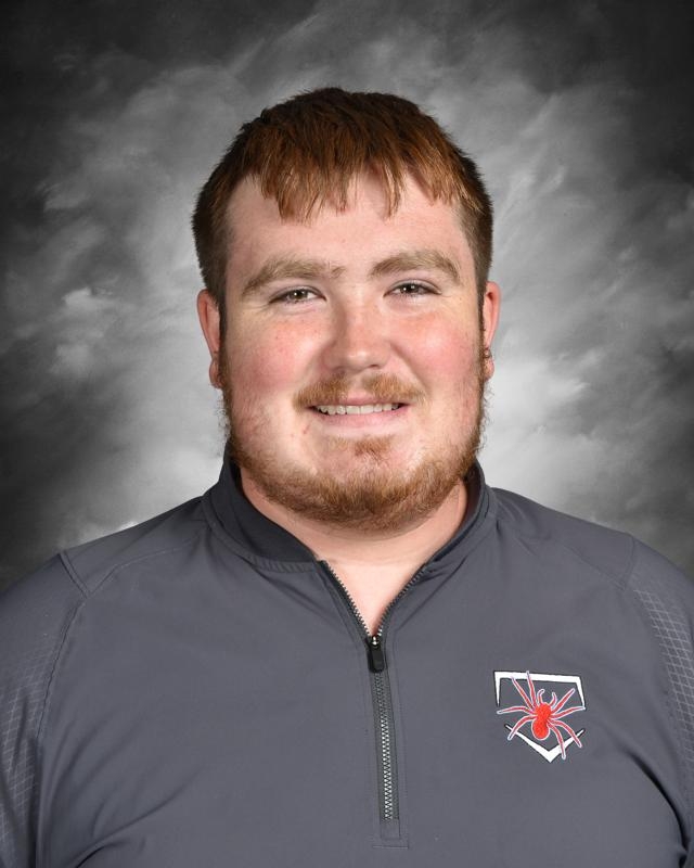 Smiling person with short hair and a beard, wearing a gray jacket with a red spider logo on a cloudy background.