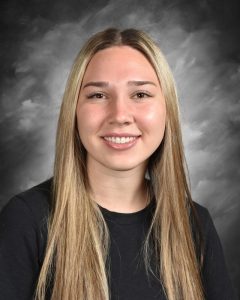 Smiling woman with long straight blonde hair, wearing a black shirt against a cloudy gray backdrop.