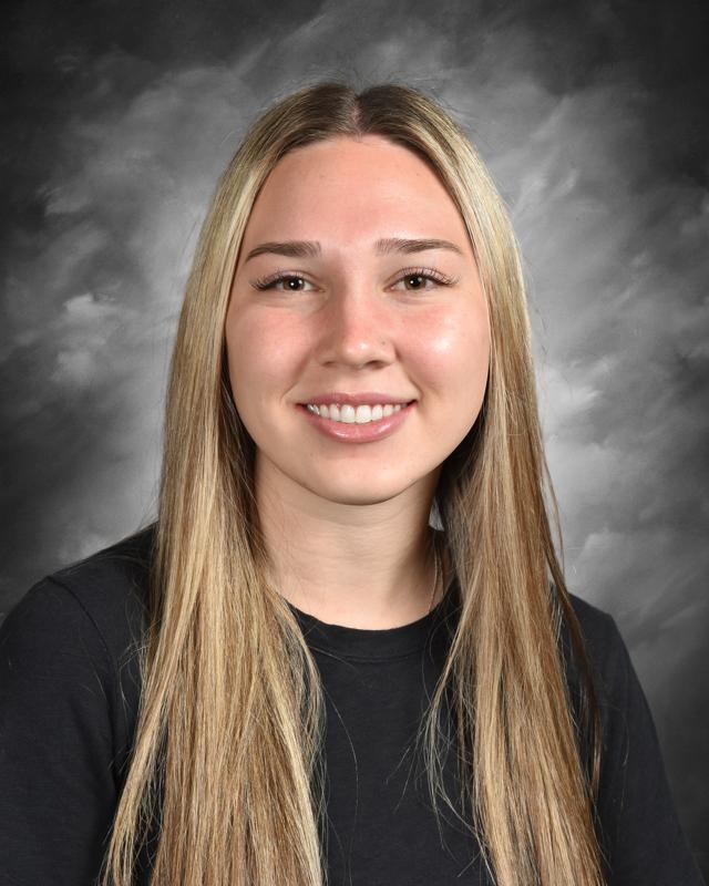Smiling woman with long straight blonde hair, wearing a black shirt against a cloudy gray backdrop.