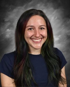 Smiling person with long, dark hair and a nose ring, wearing a dark shirt against a gray, cloudy background.