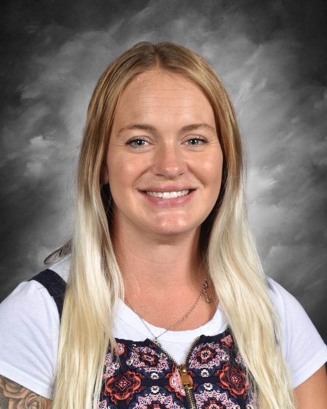Smiling woman with long blonde hair, wearing a patterned top, against a gray studio background.