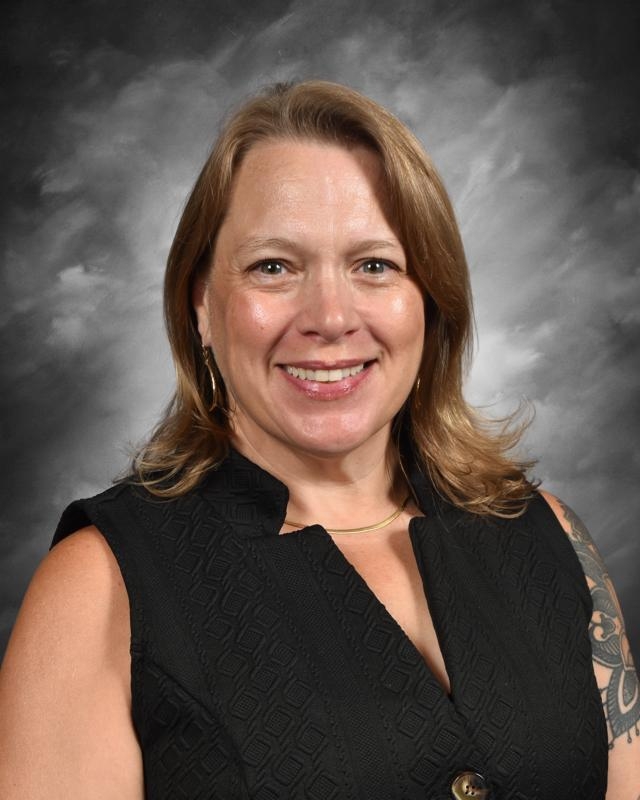 Smiling woman with shoulder-length hair in a black sleeveless top, against a gray cloudy background.