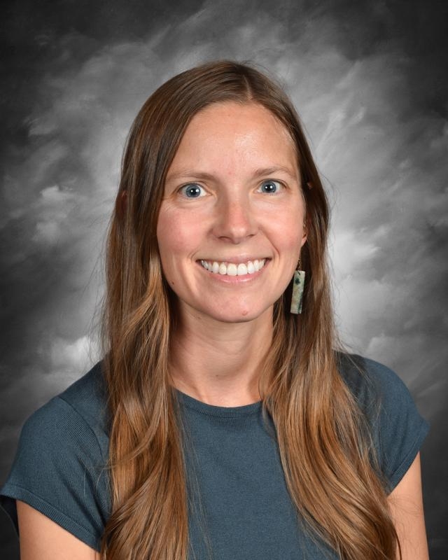 Smiling woman with long brown hair, wearing a dark shirt and earrings, against a cloudy gray background.