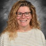 Smiling woman with curly hair and glasses, wearing a striped shirt, against a gray background.